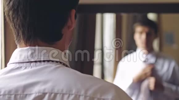 年轻的帅哥穿着白色衬衫站在镜子旁系着领带从人到镜子的焦点3840x2160视频的预览图