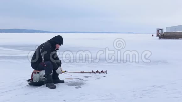 穿着深色衣服的老渔夫在冰冻的河上钓着冬天的鱼竿视频的预览图