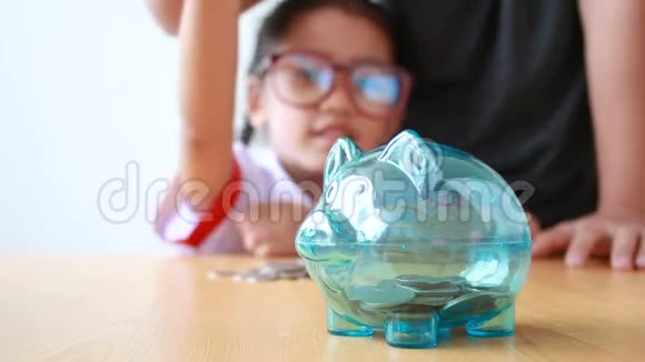 泰国幼儿园的亚洲小女孩穿着学生制服用妈妈的手把钱币放进透明的储蓄罐里选择焦点视频的预览图