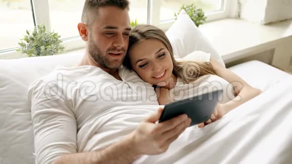 年轻可爱的一对情侣早上在卧室里躺在床上用平板电脑聊天视频的预览图