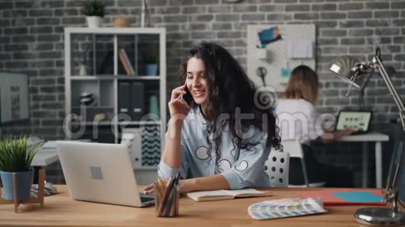 微笑的女人在办公室里用手机说话在桌子上拿着笔记本电脑视频的预览图