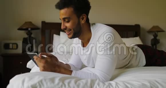 年轻人用平板电脑躺在床上快乐地微笑着西班牙男人在床上聊天视频的预览图