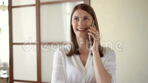 穿着白色上衣的休闲年轻女子站在客厅里笑着笑着打电话视频的预览图