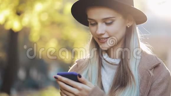 年轻女子穿着外套用智能手机站在秋天公园户外科技阳光视频的预览图