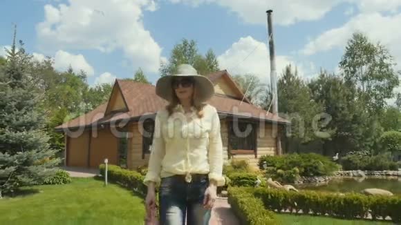 戴着帽子戴着墨镜的金发女郎走在乡间别墅的小径上视频的预览图
