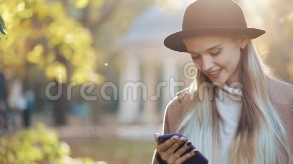 年轻女子穿着外套用智能手机站在秋天公园户外科技阳光视频的预览图