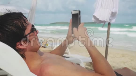 年轻人使用智能手机躺在阳光下的热带海滩与绿松石海背景3840x2160视频的预览图