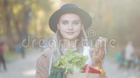 年轻漂亮的女人穿着时髦的外套站在公园里手里拿着一包产品微笑着看着视频的预览图