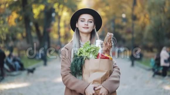 年轻漂亮的女人穿着时髦的外套站在公园里手里拿着一包产品微笑着看着视频的预览图