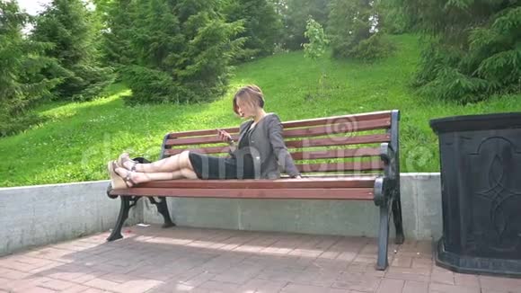 年轻时尚的女人坐在长凳上在公园里使用智能手机晴天视频的预览图