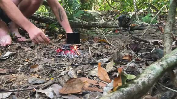 傍晚时分男子手把树枝放入热带雨林的小篝火中在篝火上做饭视频的预览图