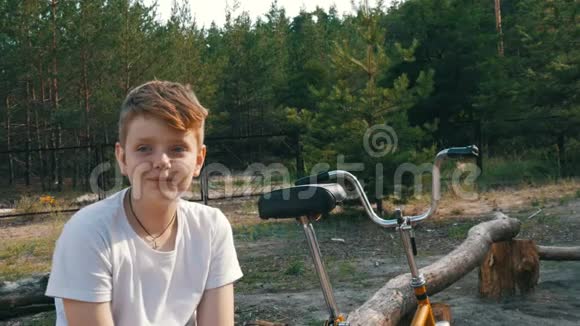 可爱的金发碧眼一个十几岁的男孩坐在自行车旁边的公园里微笑在夏天说话情感视频的预览图