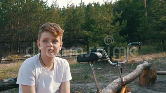 可爱的金发碧眼一个十几岁的男孩坐在自行车旁边的公园里微笑在夏天说话情感视频的预览图