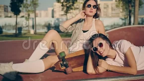 两个快乐的年轻女孩和长板一起玩同时坐在溜冰公园夏季时尚休闲视频的预览图