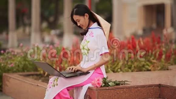 这位美丽的亚洲女孩在街上的笔记本电脑上说话穿着一件漂亮的民族服装背着一顶帽子视频的预览图
