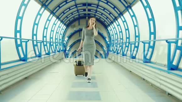 年轻的商务女性带着旅行袋到达终点站穿过玻璃隧道使用智能手机Stadicam视频的预览图