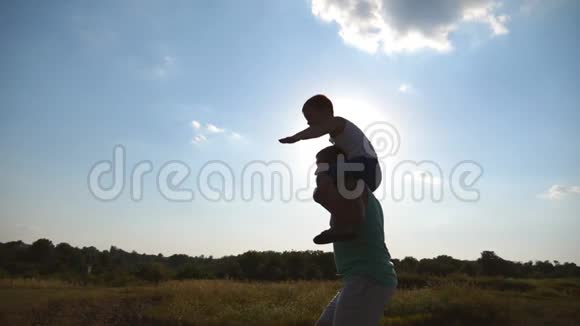 快乐的父亲和他的小儿子在夕阳下的绿野里奔跑孩子在阳光明媚的草地上坐着爸爸视频的预览图