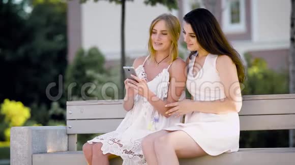 夏日阳光明媚两个穿着裙子的年轻笑女坐在长凳上休息看着照片视频的预览图