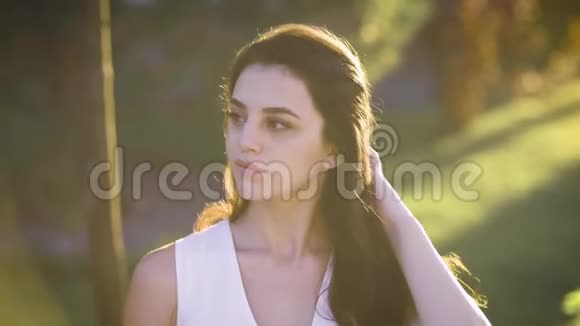 年轻迷人的微笑女人穿着白色连衣裙坐在公园的贝恩奇上在光线下闪耀着美丽的光芒视频的预览图