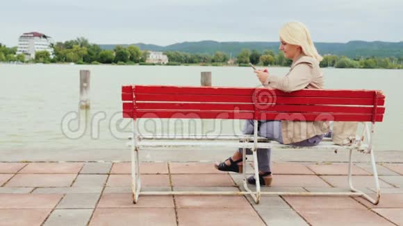 女人坐在一个大湖背景上的长凳上使用手机匈牙利巴拉顿湖视频的预览图