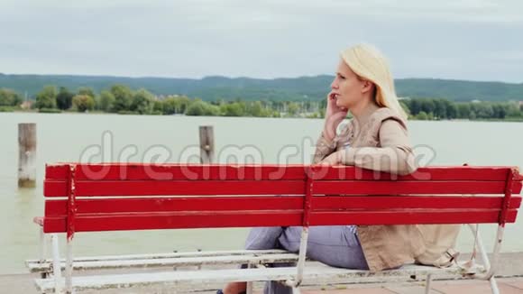 女人坐在一个大湖背景上的长凳上在电话里说话匈牙利巴拉顿湖视频的预览图