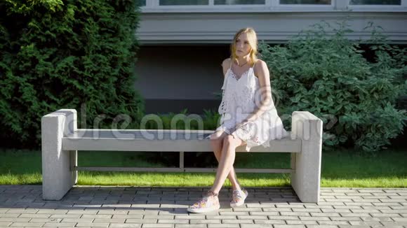 夏天这位可爱的年轻女子坐在一条长凳上穿着白色的蕾丝连衣裙想着并喜欢独处视频的预览图