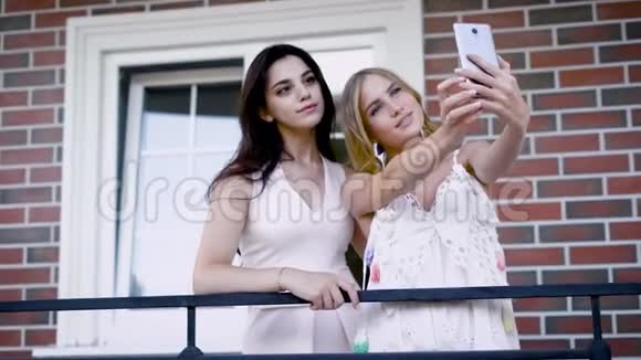 两位穿着白色连衣裙的美女正在智能手机上以砖墙和窗户为背景自拍视频的预览图