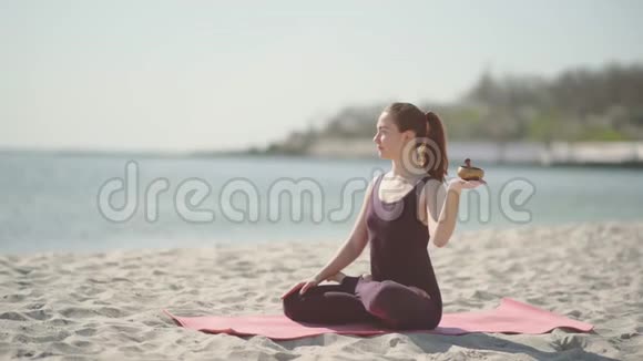 阳光明媚的一天年轻漂亮的女人在海滩上练习瑜伽女人用藏式唱碗冥想视频的预览图