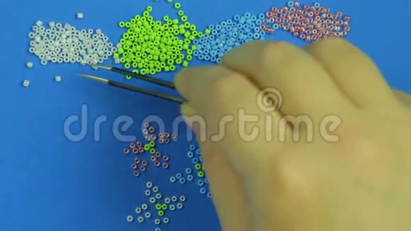 蓝色背景上的雌性手用镊子从多种颜色的珠子中抽出花朵特写视频的预览图