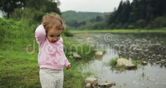 金发卷曲的小女孩在河里扔石头山脉背景视频的预览图