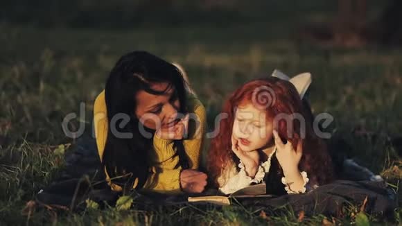 美丽的妈妈和她可爱的小女儿躺在公园的草地上看书他们说得很好快乐视频的预览图