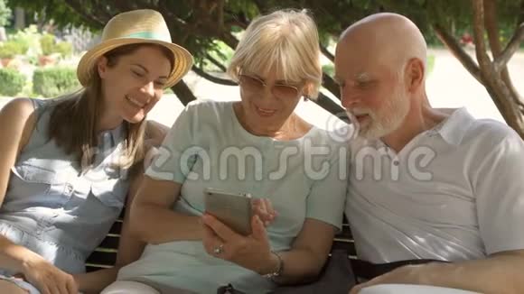 快乐的家庭享受假期老年人和他们的女儿坐在公园里浏览智能手机视频的预览图
