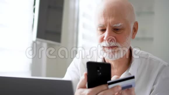 英俊的胡子老人坐在家里使用智能手机信用卡在线购物视频的预览图