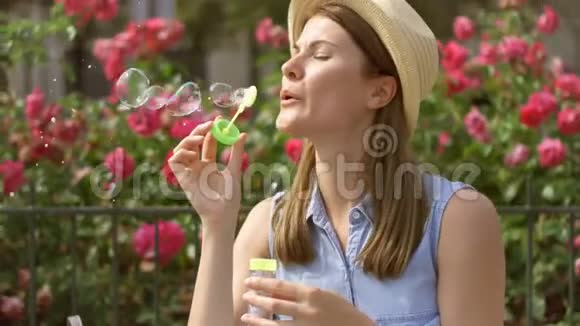夏日阳光明媚年轻有趣的女孩吹着肥皂泡背景上美丽的粉红色花朵视频的预览图