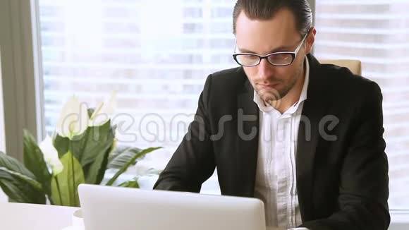 英俊的商人戴着笔记本电脑上的眼镜对着镜头微笑视频的预览图