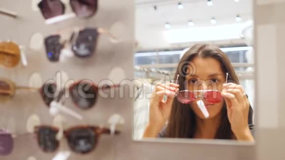 年轻迷人的混合种族女孩尝试太阳镜附近的镜子购物中心时髦的嬉皮士时装看起来很慢视频的预览图