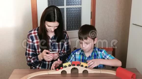 一起玩妈妈看着电话儿子正在玩一条木制铁路火车马车和隧道坐在那里视频的预览图