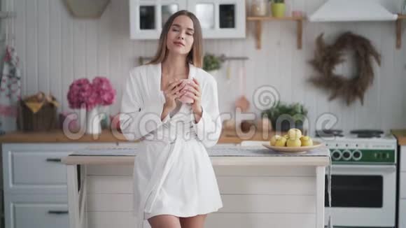 穿着白色浴袍在厨房喝咖啡或茶的漂亮女孩的肖像视频的预览图