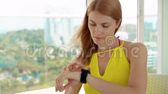 微笑快乐的女人穿着黄色的裙子坐在阳台上利用她的智能手表和朋友聊天视频的预览图