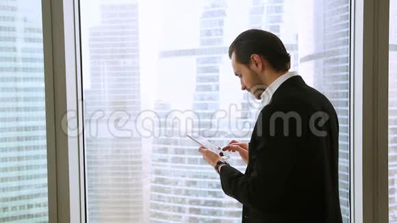 自信快乐的商人站在办公室窗口使用平板智能手机视频的预览图