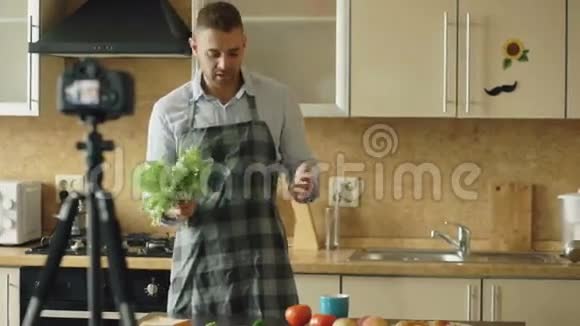 穿围裙的年轻帅哥在厨房里用dslr摄像机拍摄有关烹饪的视频食物博客视频的预览图