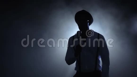 音乐家在烟雾中的白光中唱着麦克风黑色背景剪影慢动作视频的预览图
