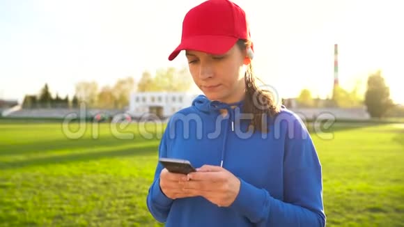拥有无线耳机和智能手机的女人选择音乐并在日落时穿过体育场慢动作视频的预览图