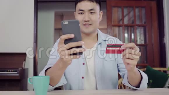 亚裔男子使用智能手机进行网上购物和信用卡在互联网上的客厅家视频的预览图