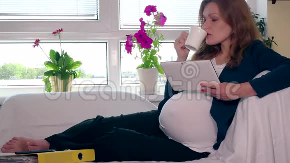 快乐的孕妇喝茶用平板电脑在沙发上放松视频的预览图