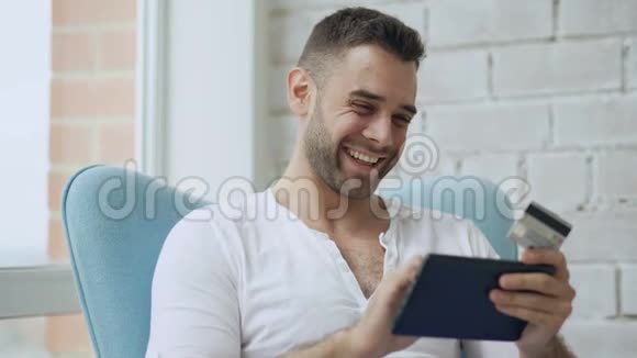 坐在家阳台上用数字平板电脑上网购物的微笑青年视频的预览图