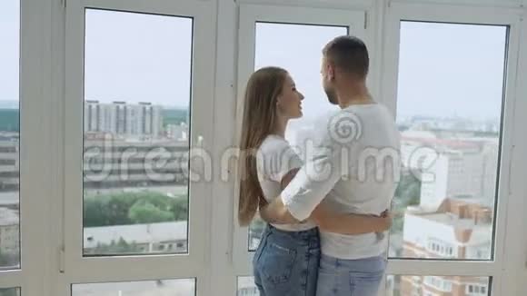 年轻的幸福夫妇拥抱站在窗户附近享受新公寓的景色视频的预览图
