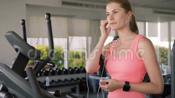 适合积极运动的女人在天鹅绒模拟器上做运动使用她的智能手机用耳机听音乐视频的预览图