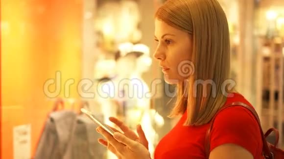 漂亮迷人的年轻女子靠近商场的橱窗商店检查她的智能手机的商店价格视频的预览图