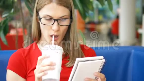聪明的口渴学生戴着眼镜坐在咖啡馆里喝着纸杯里的可乐读着她的笔记视频的预览图